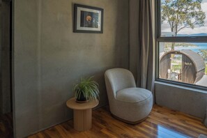 A room with a wooden floor, a grey armchair, a small round table with a plant, a framed picture on the wall, and a view of a wooden structure outside through a window. - Mabels Lookout - Sauna| Bath| Sunsets| Views| Fire (North Bruny)