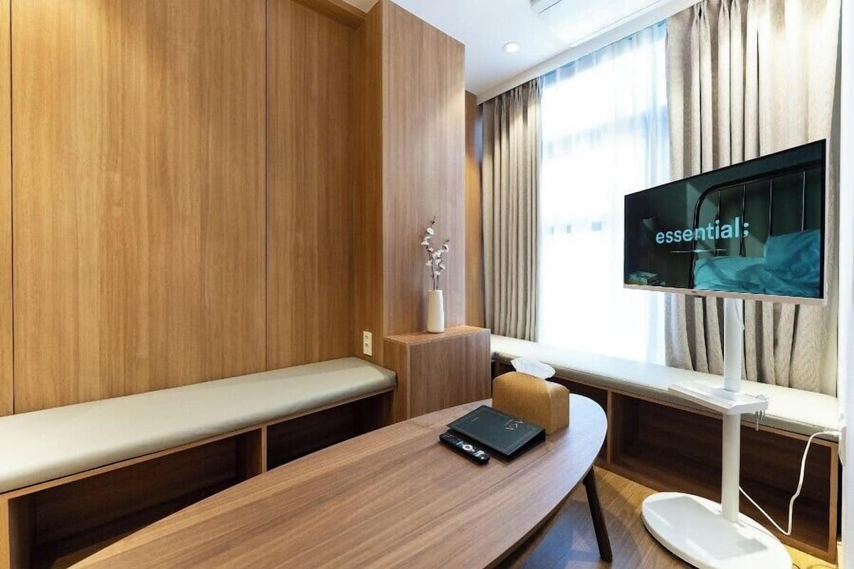 Ein modernes Hotelzimmer mit Holzvertäfelung, einem großen Fenster mit Vorhängen, einem Flachbildfernseher, einem Holztisch mit einem Laptop und einer Bank.