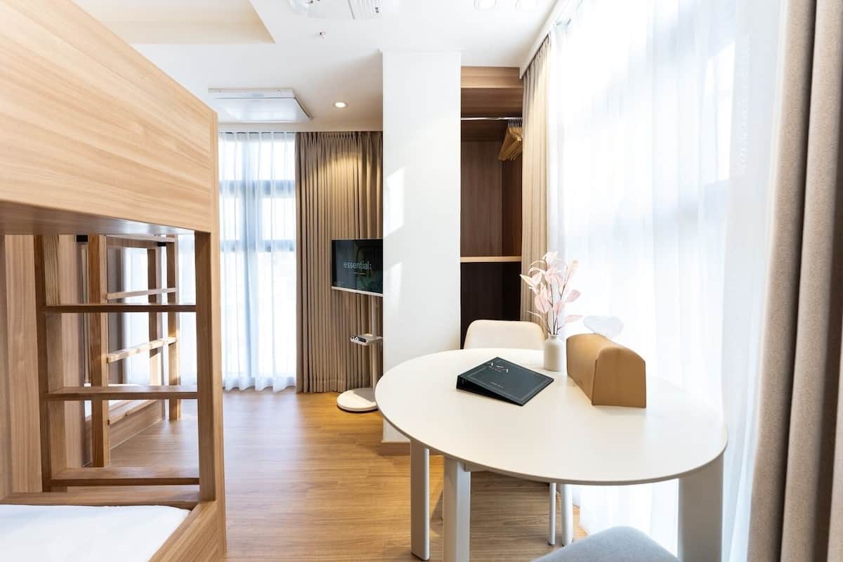 Ein modernes Hotelzimmer mit Holzboden, einem runden weißen Tisch mit einem Buch, einer beigen Couch, einem Fernseher an der Wand und einem Kleiderschrank mit Kleidung.