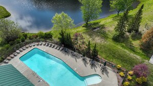 En swimmingpool med en omgivande terrass, utsikt över en sjö och ett grönområde med träd och buskar.