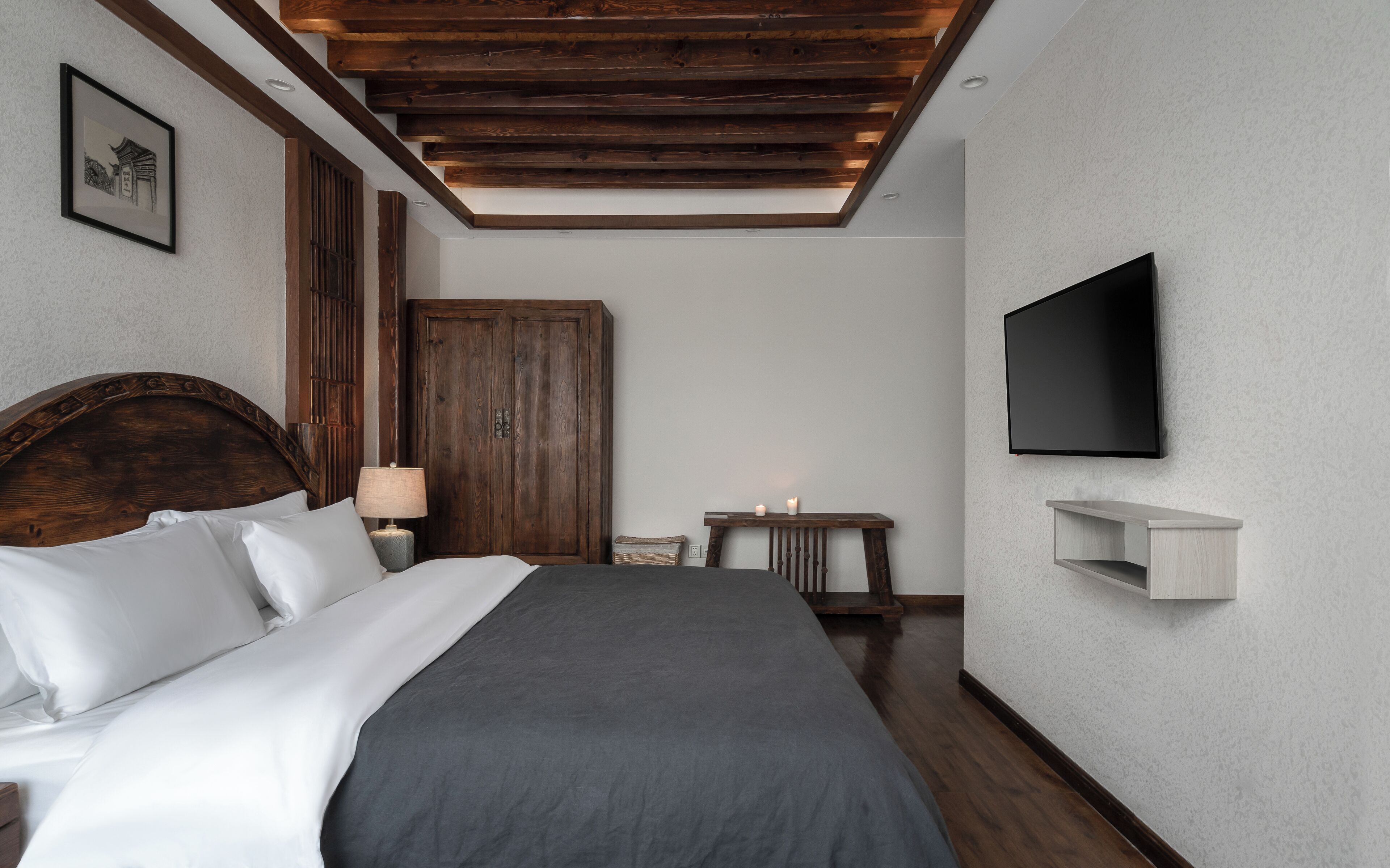 A bedroom with a wooden headboard, a bed with white and gray bedding, a wooden wardrobe, a bedside table with a lamp, a small table with candles, and a mounted TV on the wall.