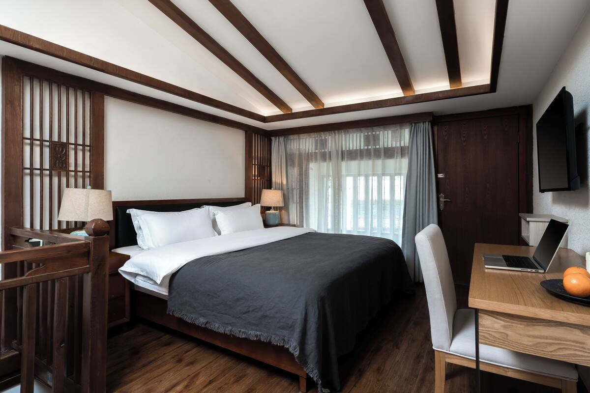 Habitación de hotel con una cama de madera, un escritorio con una computadora portátil, una silla y un televisor fijado en la pared.