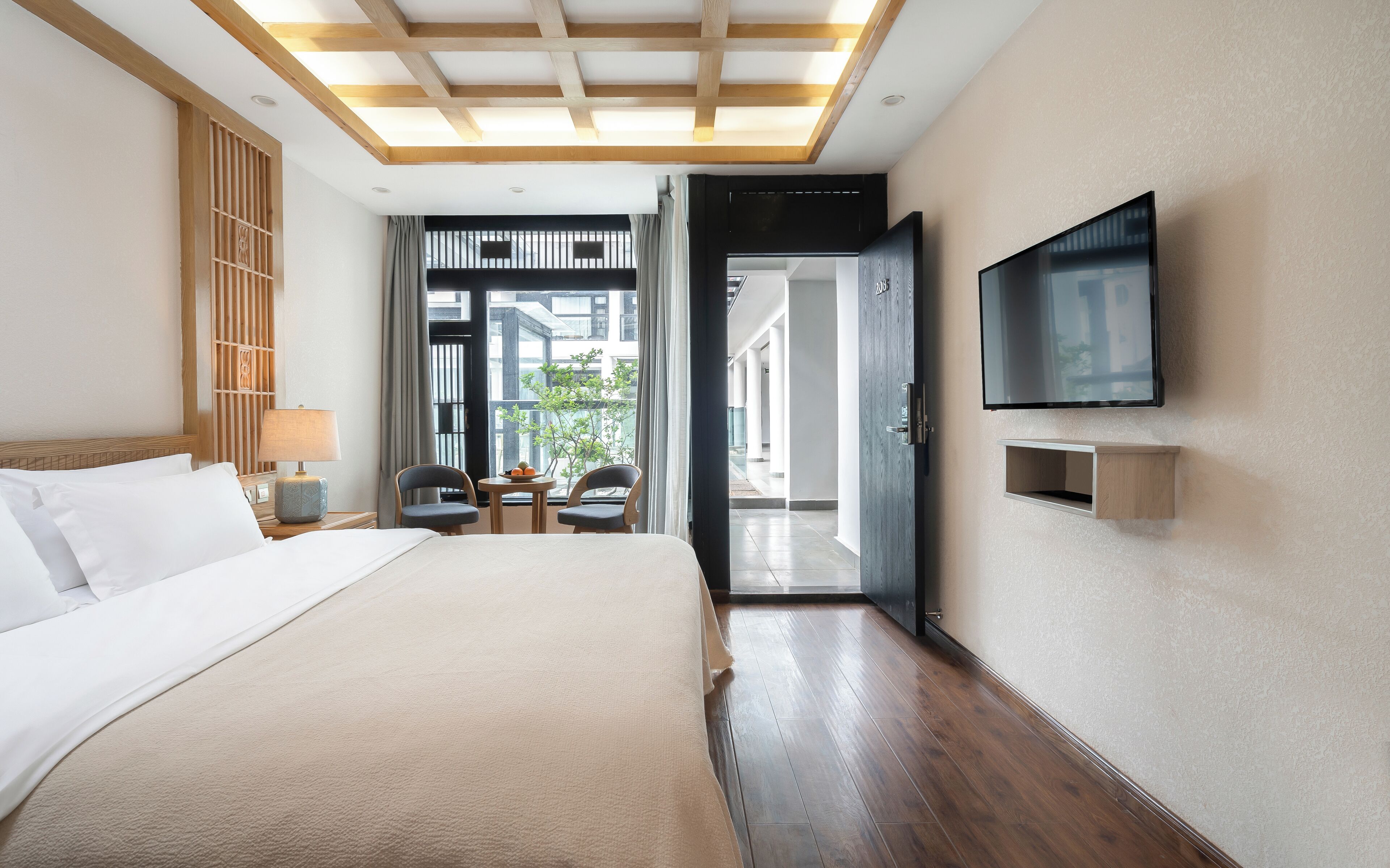A hotel room with a bed, a TV mounted on the wall, a small table with chairs, and a view through an open door.