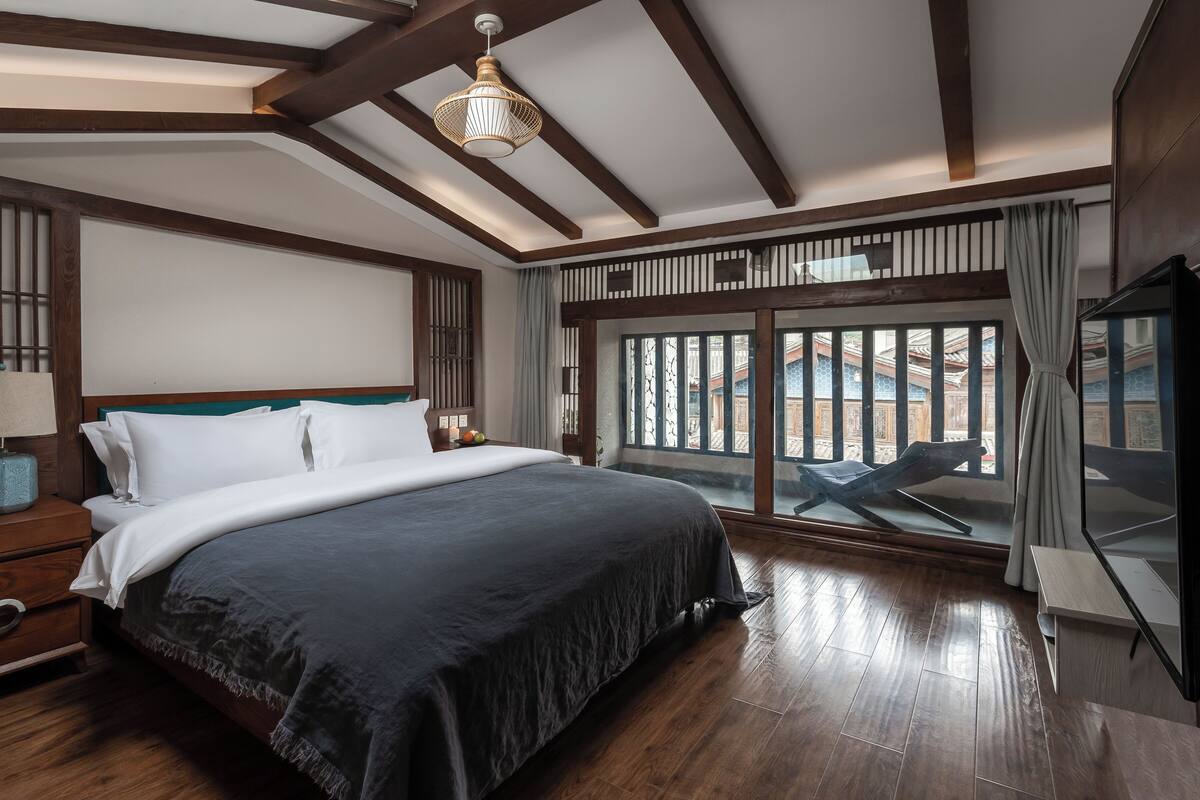 Un dormitorio con una cama grande, vigas de madera, una mesita de noche con una lámpara, un ventanal con vistas y un televisor.