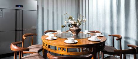 A dining area with a round table, chairs, a vase with flowers, and a refrigerator in the background.