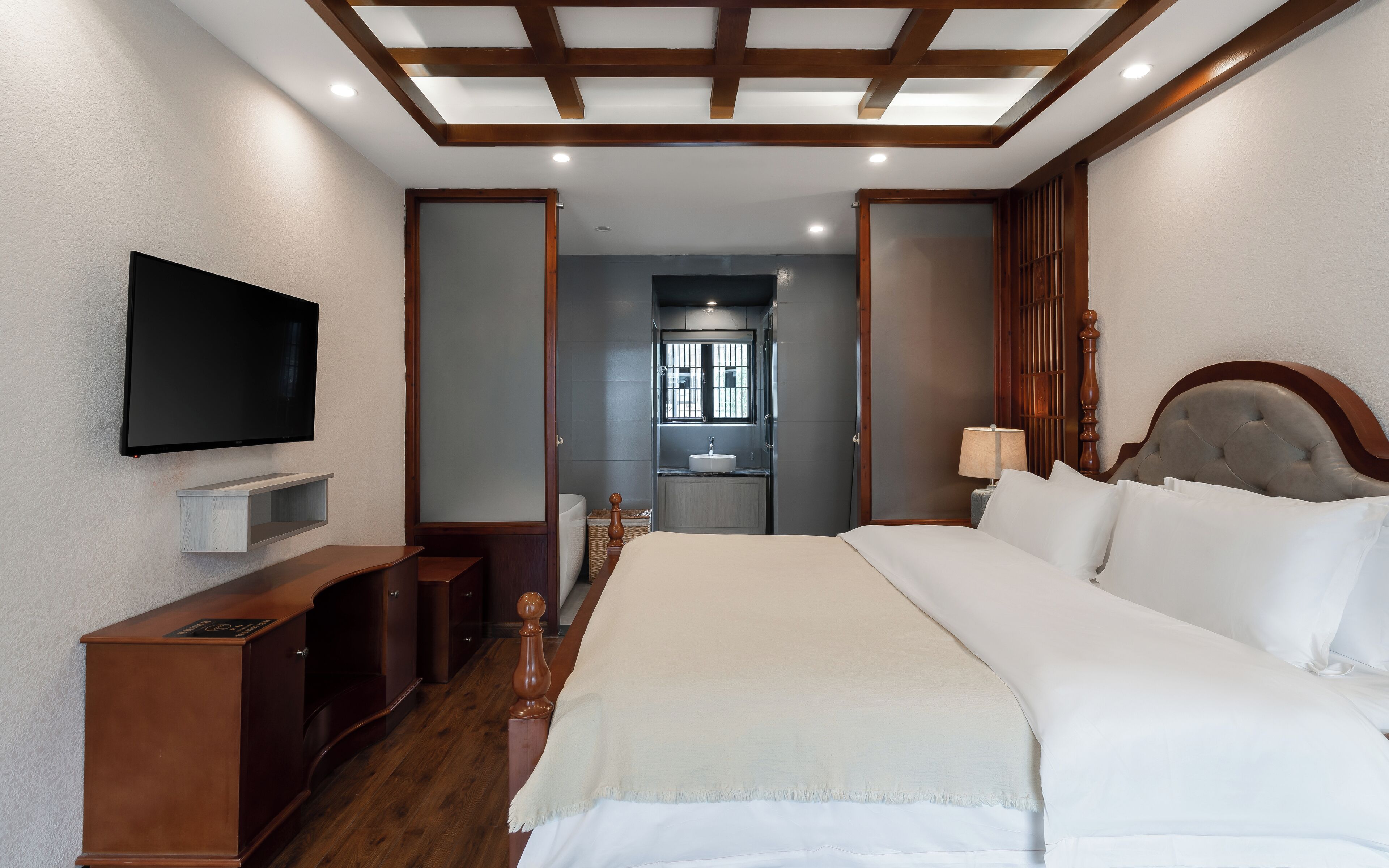 A hotel room with a bed, a TV mounted on the wall, a wooden nightstand, and a bathroom visible through an open door.