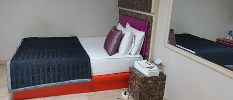 A bed with a black cover, a red base, and a headboard with a purple pillow. There is a small table with a mirror and a stack of white towels.