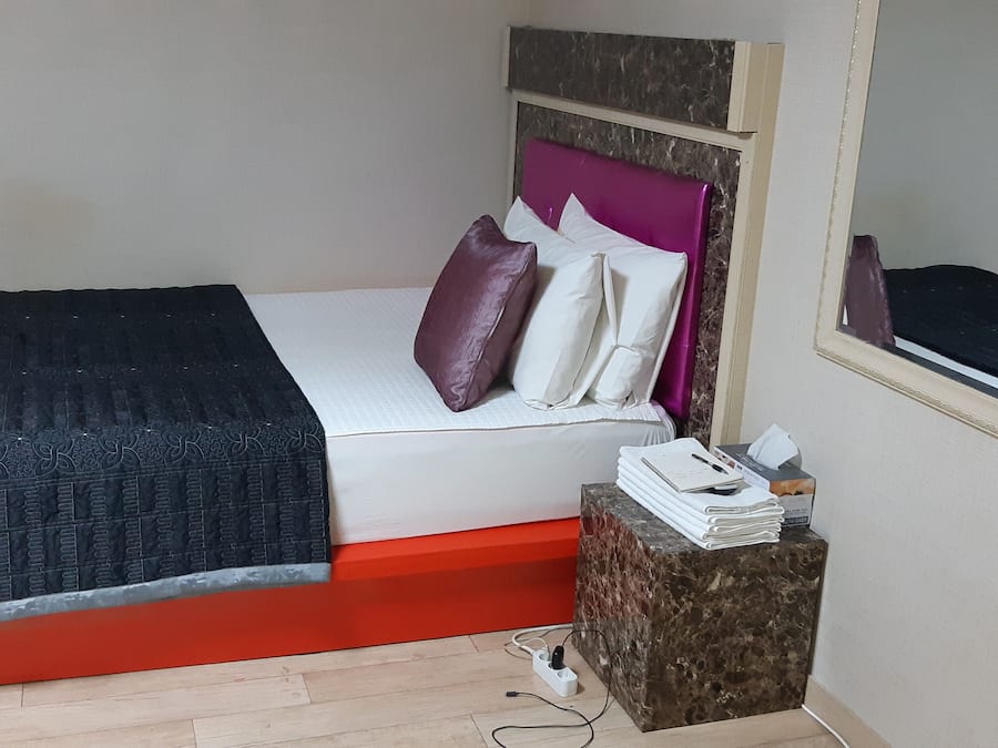 A bed with a black cover, a red base, and a headboard with a purple pillow. There is a small table with a mirror and a stack of white towels.