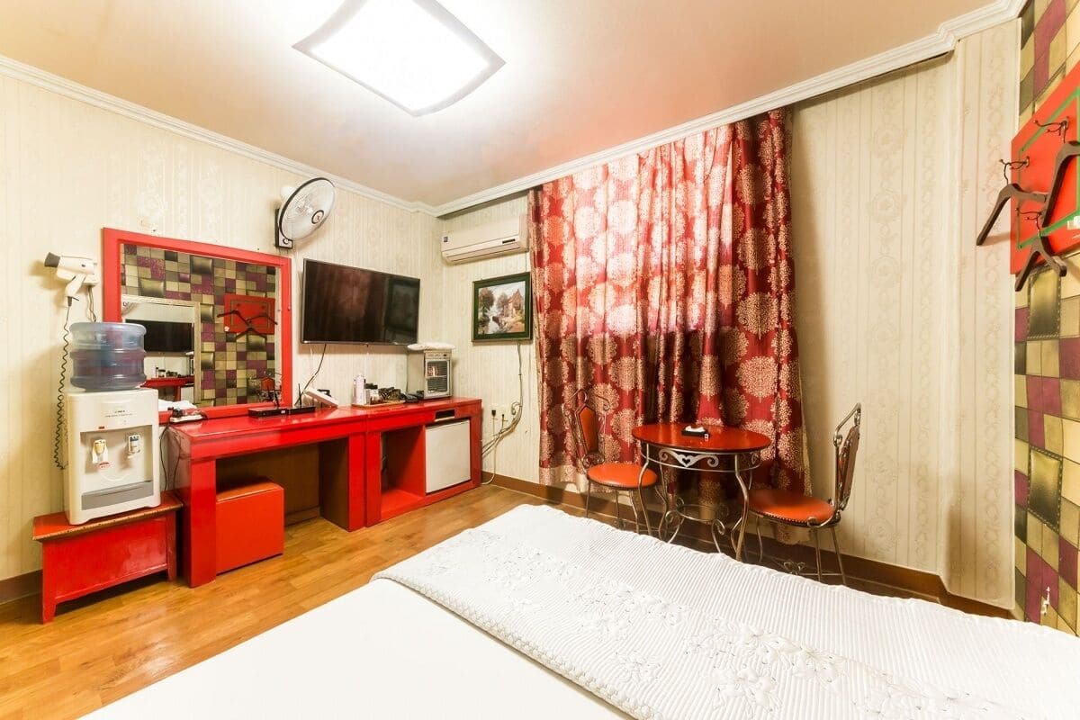 A compact hotel room with a bed, a desk with a computer, a small table with chairs, a red cabinet, and a large mirror.