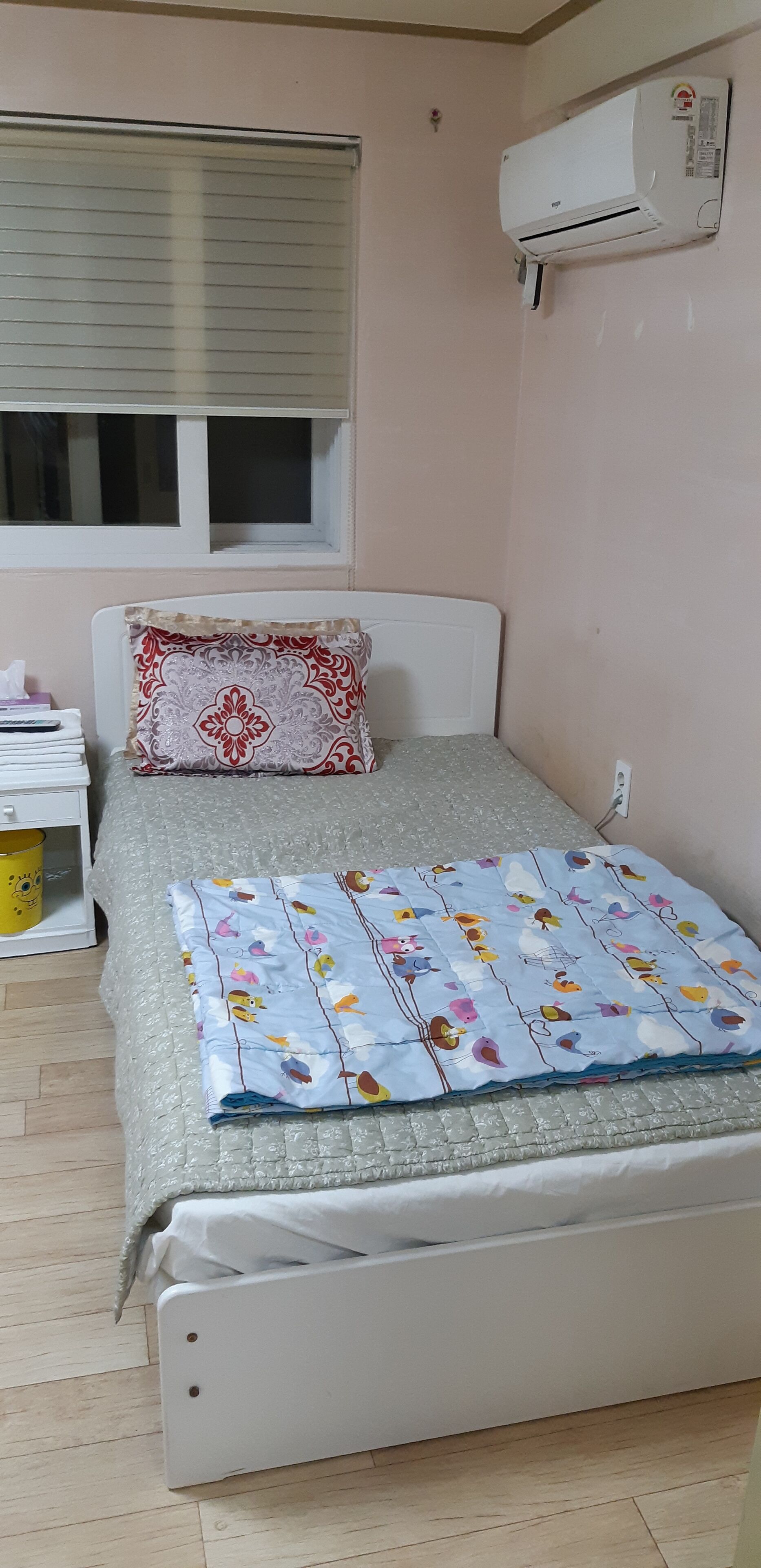 A small, neatly arranged bedroom with a bed, a pillow, a bedside table with a lamp, and an air conditioning unit mounted on the wall.