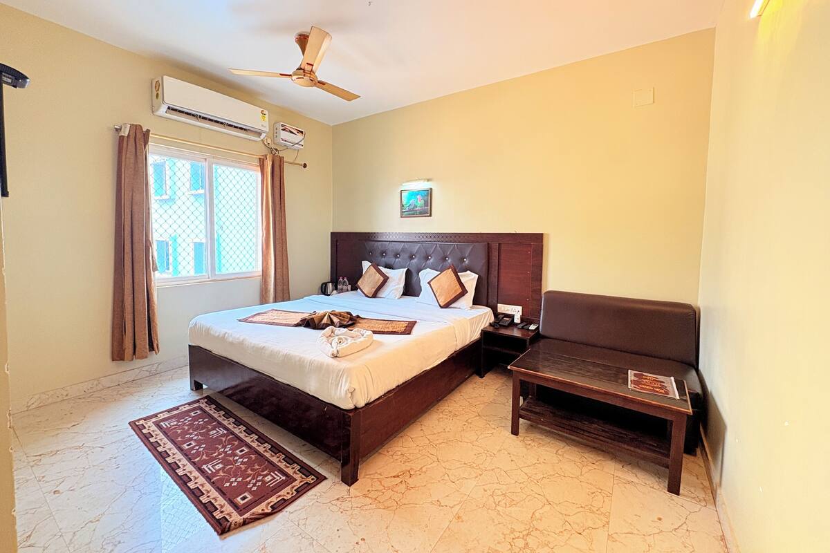 A hotel room with a bed, a nightstand, a chair, a ceiling fan, an air conditioning unit, a window with curtains, and a rug.