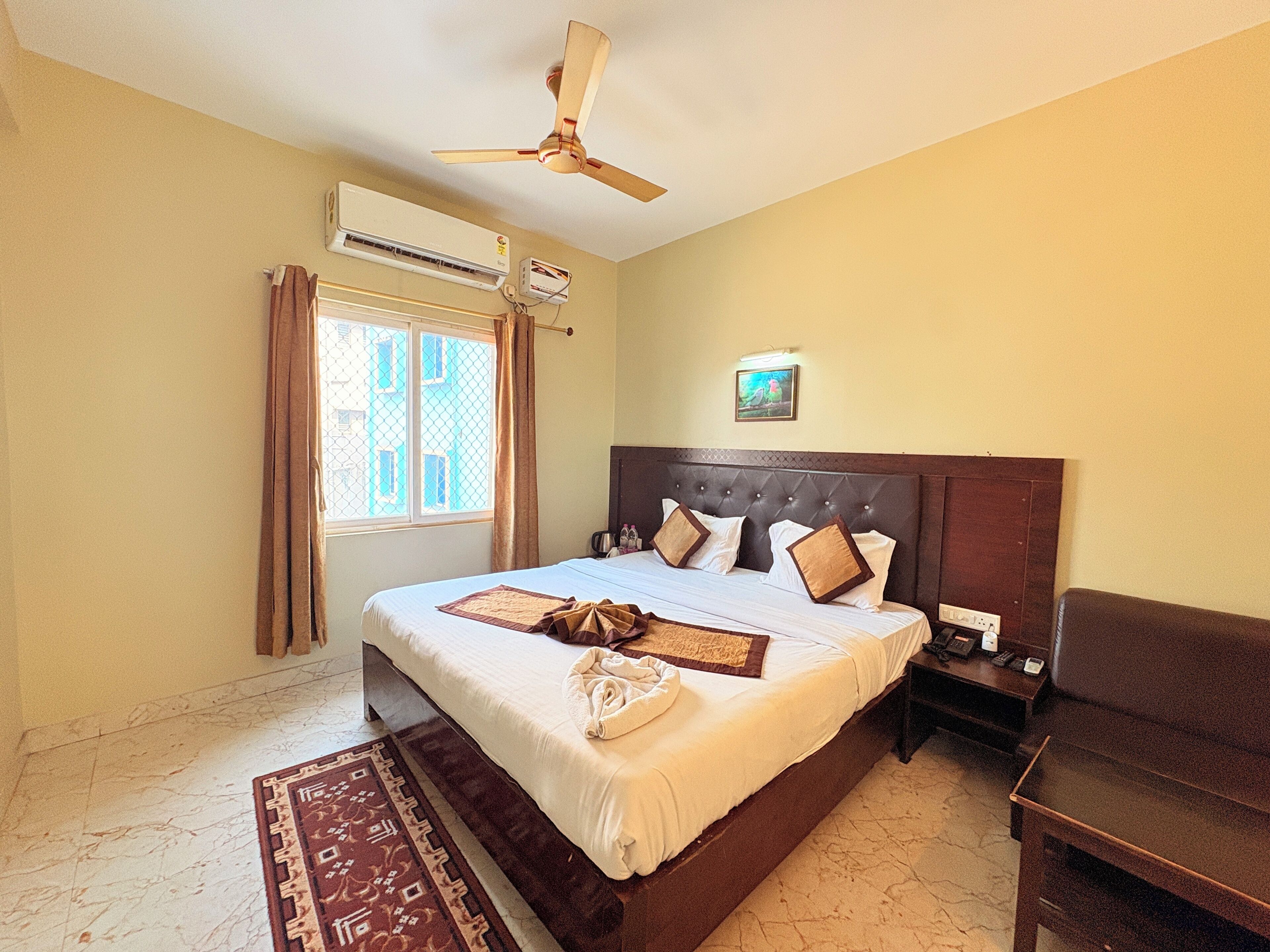 A hotel room with a large bed, a ceiling fan, an air conditioning unit, a window with curtains, and a bedside table with a phone and a lamp.