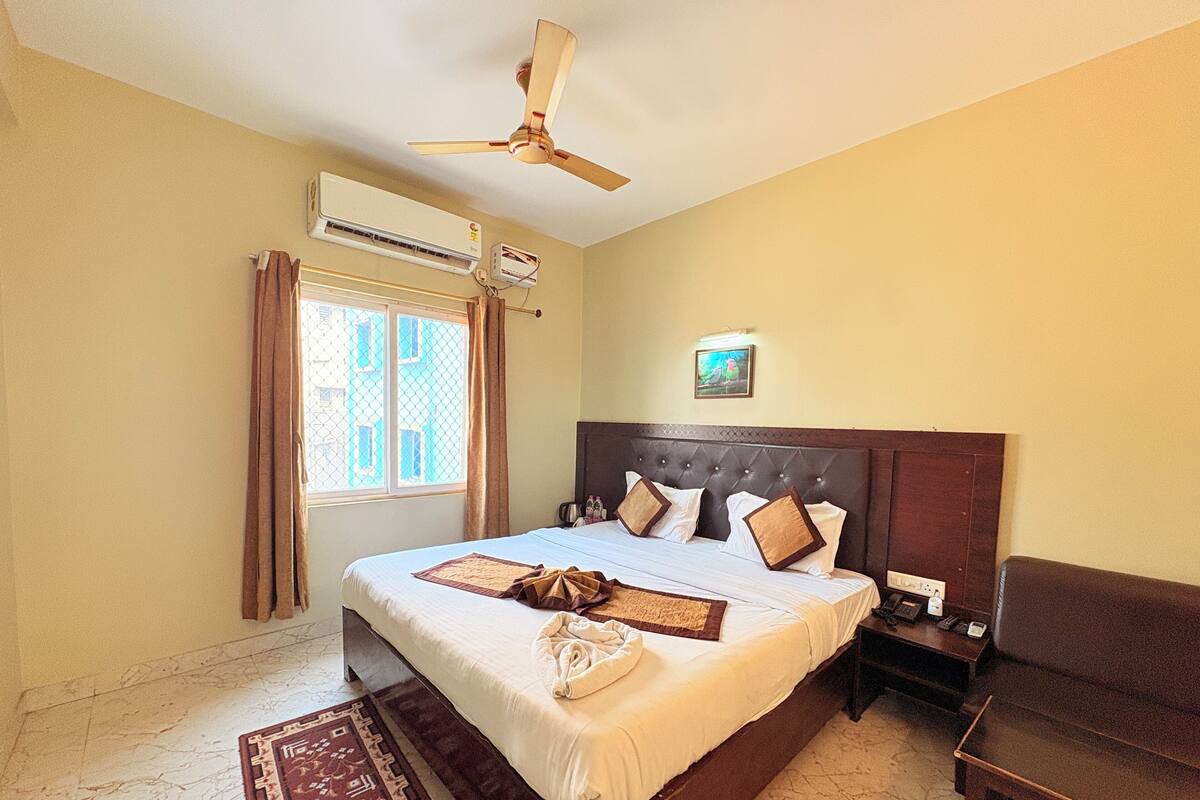 A hotel room with a large bed, a ceiling fan, an air conditioning unit, a window with curtains, and a bedside table with a phone and a lamp.