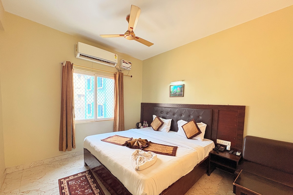 A hotel room with a large bed, a ceiling fan, an air conditioning unit, a window with curtains, and a bedside table with a phone and a lamp.