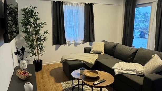 A modern living room with a sectional sofa, a coffee table, a potted plant, and a flat-screen TV mounted on the wall. - Beautiful vacation apartment in the Altmark (Goldbeck)