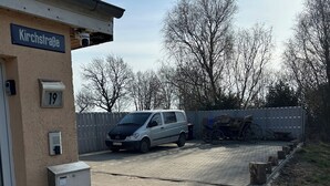 A building with a sign reading 'Kirchstraße 19,' a parked car, and a paved area with a fence and trees in the background. - Beautiful vacation apartment in the Altmark (Goldbeck)