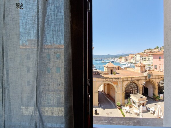 Vue sur une ville côtière depuis une fenêtre, avec un port, des bâtiments et une rue où sont installées des terrasses.