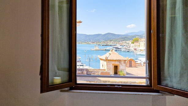 A view through an open window of a coastal town with boats, a small building, and mountains in the distance. - Porta A Mare Home Apartment with Air Conditioning (Portoferraio)