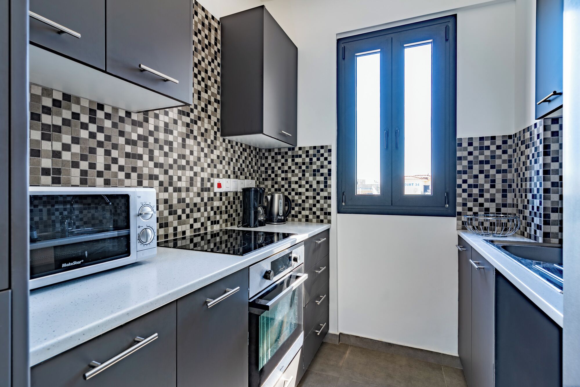 Cozinha compacta com micro-ondas, fogão e chaleira. Há uma janela com vista para edifícios.