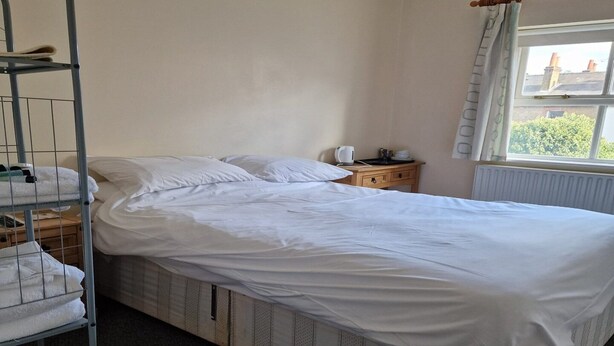 Ein Einzelzimmer mit einem Metallstockbett, einem Holznachttisch, einem Wasserkocher und einem Fenster mit Vorhängen.