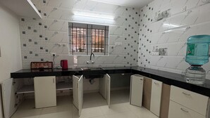 A modern kitchen with white marble tiles, a black countertop, and a window with metal bars. - BBR Holiday Home (Ooty)