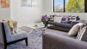 A modern living room with a grey sofa, a grey armchair, and a coffee table. There are three framed artworks on the wall and a window with a view of trees. - Parkside Elegance on Puoro St Near Shopping Centre (Auckland)