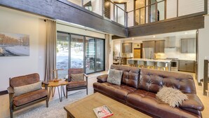 Un vaste espace de vie agrémenté d’un canapé en cuir marron, d’une table basse en bois et d’un tableau accroché au mur. Un escalier relie l’espace de vie à un niveau supérieur.