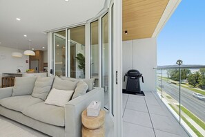 A modern living room with a sofa, a grill, and a view of a road and greenery. - Kailani 302: Luxury Waterfront, Bay & Marina Views (Batemans Bay)