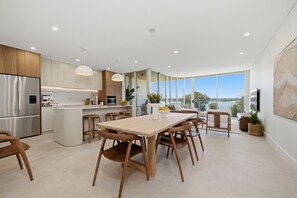 A modern kitchen with a large island, stainless steel appliances, and a dining area with wooden chairs. - Kailani 302: Luxury Waterfront, Bay & Marina Views (Batemans Bay)