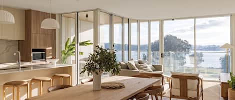 Une cuisine moderne dotée d’un îlot central, d’un coin repas avec une table et des chaises en bois, et d’une grande porte-fenêtre coulissante donnant sur un balcon avec vue sur l’eau.