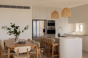 A modern kitchen with white cabinetry, a central island, and a dining area with a wooden table and chairs. - Kailani 301: Designer Waterfront Apartment (Batemans Bay)
