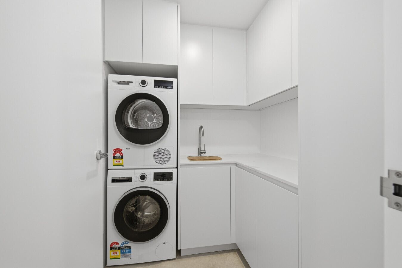 A compact laundry room with two stacked washing machines, a small sink, and a wooden cutting board.