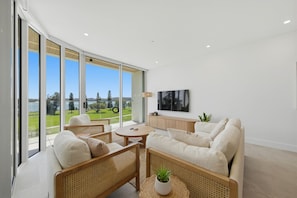 A modern living room with a large window, a flat-screen TV, and a view of a golf course. - Kailani 202: Uninterrupted Bay & Marina Views (Batemans Bay)
