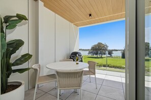 A balcony with a round table and wicker chairs, overlooking a lake and grassy area. - Kailani 202: Uninterrupted Bay & Marina Views (Batemans Bay)
