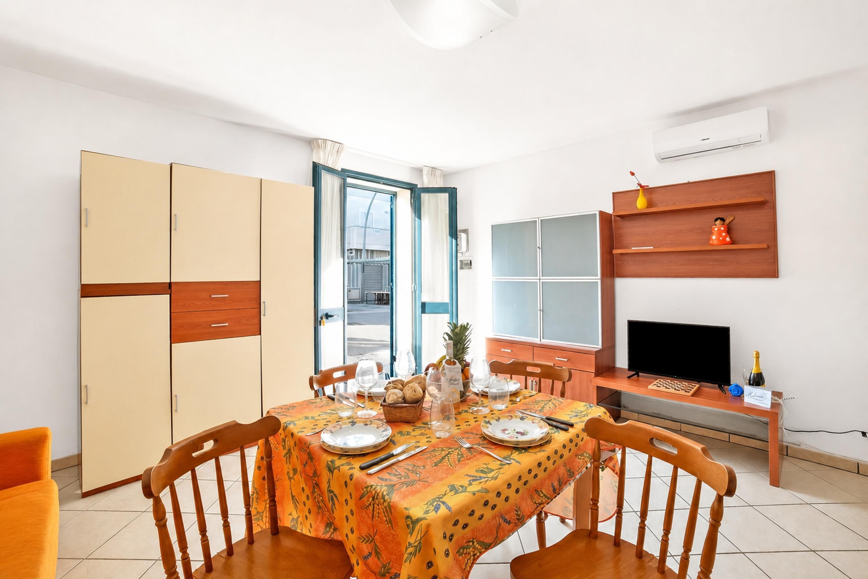 Un comedor con una mesa dispuesta para dos, un televisor sobre un mueble bajo y un armario con varias puertas.