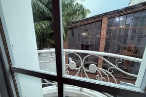 Blick durch ein Fenster mit schmiedeeisernem Geländer, ein Backsteinhaus und im Hintergrund Palmen.