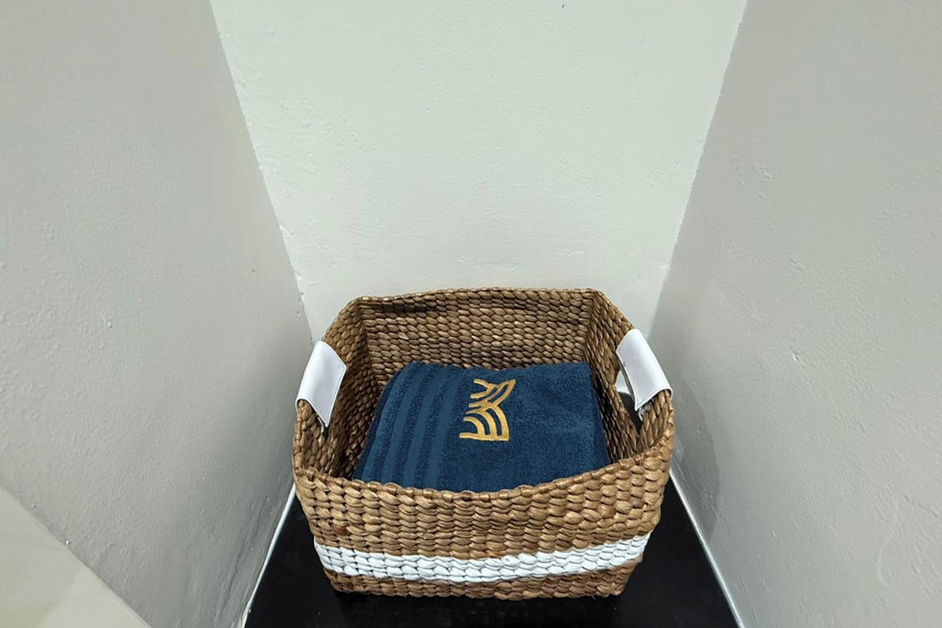 A woven basket with a blue towel and a gold emblem, placed against a plain wall.
