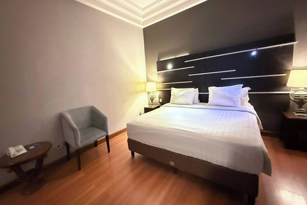 A hotel room with a large bed, a small armchair, a bedside table, and a nightstand with a lamp.