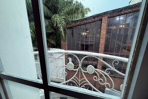 Blick aus einem Fenster mit dekorativen Metallgeländern, ein Ziegelgebäude mit großen Fenstern und eine Palme im Hintergrund.