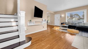Een moderne woonkamer met een houten trap, een flatscreen televisie aan de muur, een zithoek en een groot raam met jaloezieën.