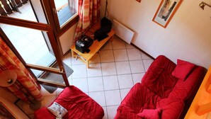 A room with a red sofa, a small wooden table with a speaker, and a window with curtains. - Duplex in Les Carroz (Araches-la-Frasse)