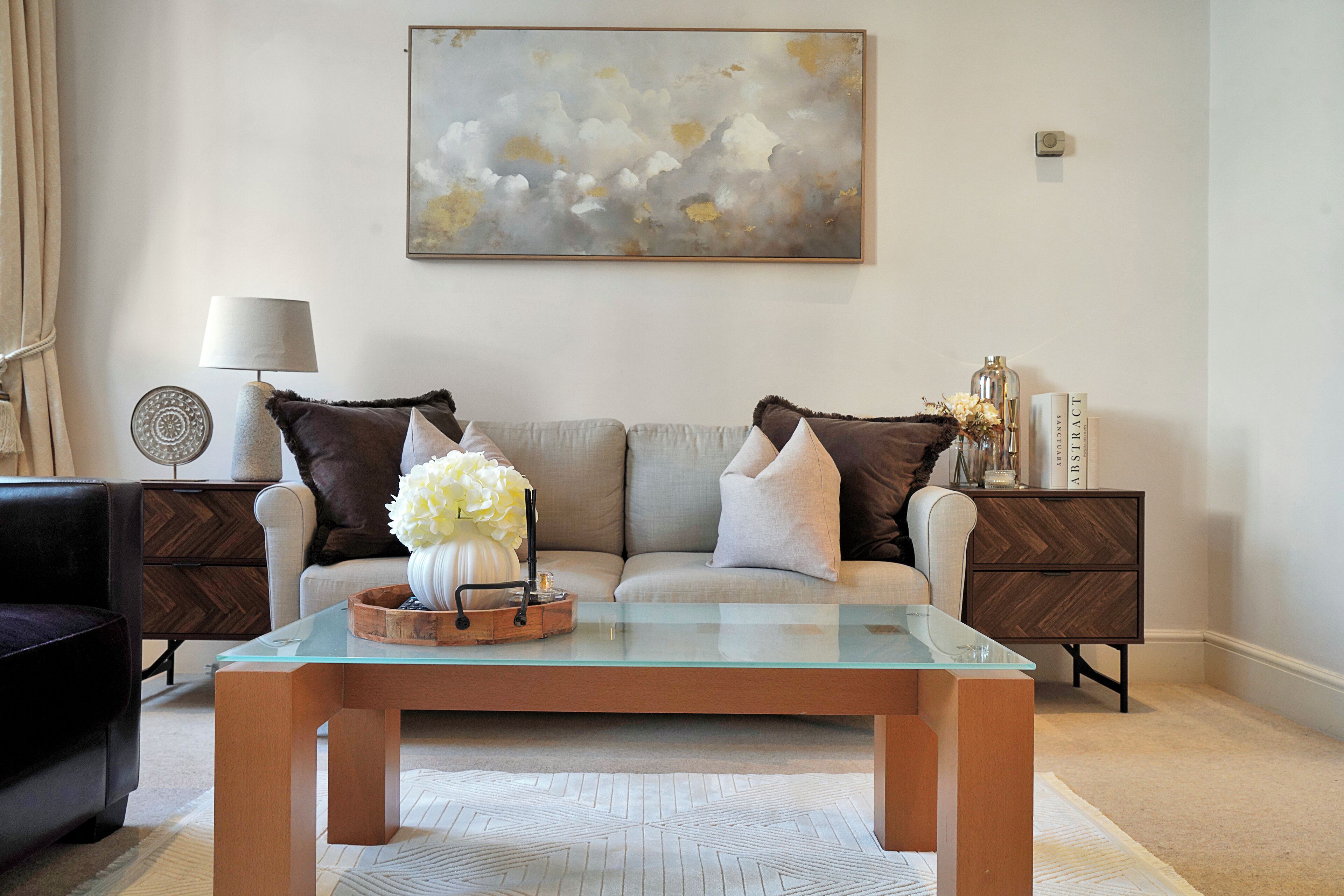 A modern living room with a glass coffee table, a sofa with decorative pillows, a side table with a lamp, and a wall-mounted abstract painting.