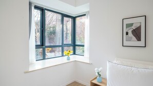 A corner bedroom with a bay window, a framed picture on the wall, and a bedside table with a vase.