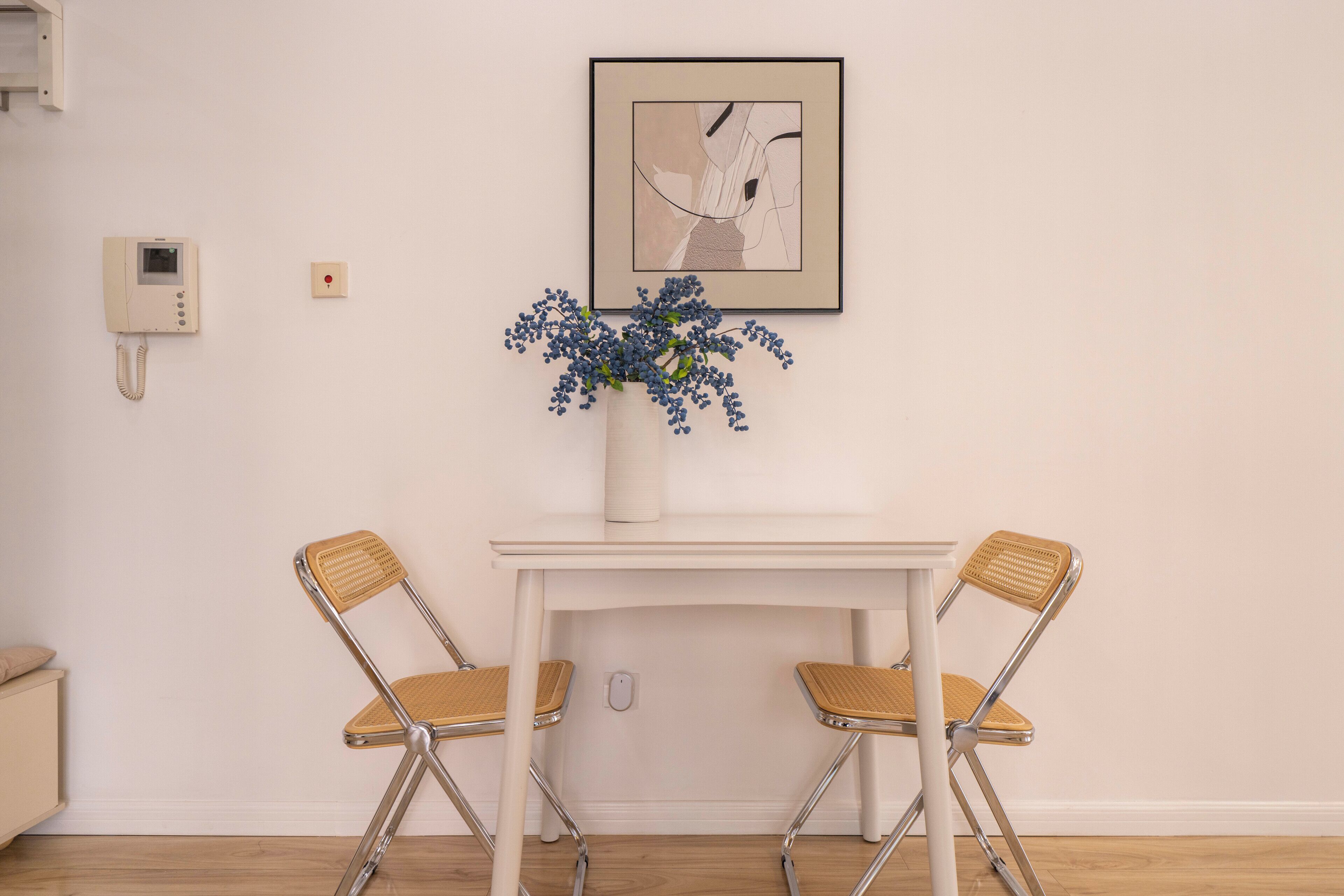 A dining area with a white table, two wicker chairs, a vase with blue flowers, a framed abstract artwork, and a wall-mounted intercom.
