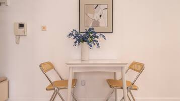 A dining area with a white table, two wicker chairs, a vase with blue flowers, a framed abstract artwork, and a wall-mounted intercom.