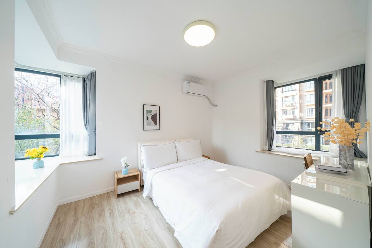 A bedroom with a bed, bedside tables, a nightstand, a window with curtains, a wall-mounted air conditioner, and a framed picture.