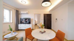 A modern living room with a round dining table, orange chairs, a white sofa, a TV, and a decorative vase. - Tyms Tianshan 2BR near Riverside Park (Shanghai)