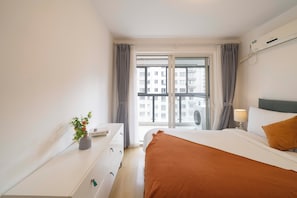 A bedroom with a bed, a dresser, a bedside table with a lamp, a window with curtains, and an air conditioning unit. - Tyms Tianshan 2BR near Riverside Park (Shanghai)