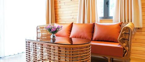 A cozy room with a wicker sofa and a round table, a window with curtains, and wooden walls.