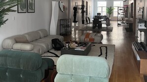 Amplia sala de estar con un sofá modular, un sillón verde y una mesa de centro. Se observan tuberías en el techo y una ventana al fondo.